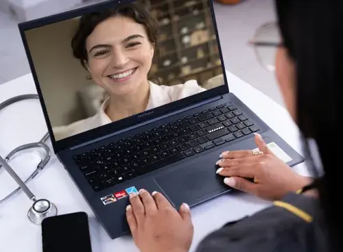 Persona frente a un portátil participando en una reunión virtual; sobre la mesa hay un celular y un estetoscopio.